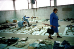 As forensic anthropologists Ewa Klonowski and Piotr Drukier work in the background, a woman walks among the rows of remains laid out with the clothing found with each skeleton during the exhumation of mass graves. Relatives looking for a loved one often recognize a piece of clothing first; a formal identification is made through forensic data and DNA samples. Thousands of Bosnians -- the majority of them Muslims -- remain missing years after the end of the war. October 2000.