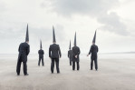 six Conemen in black suits stand on an empty beachwith their back to the viewer