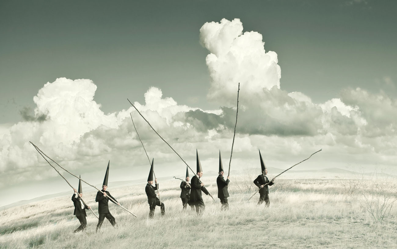 Several Conemen wander across an alien curved green landscape with big billious clouds in the sky. They each carry a long wooden pole.