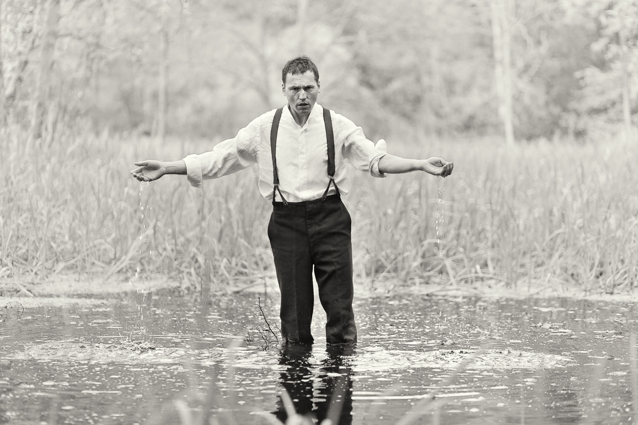 a man stands in a lake like a preacher, his arms held out, while water runs through his hands as if in baptism
