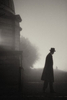 man in long coat and bowler hat looks down a road into the mist at night