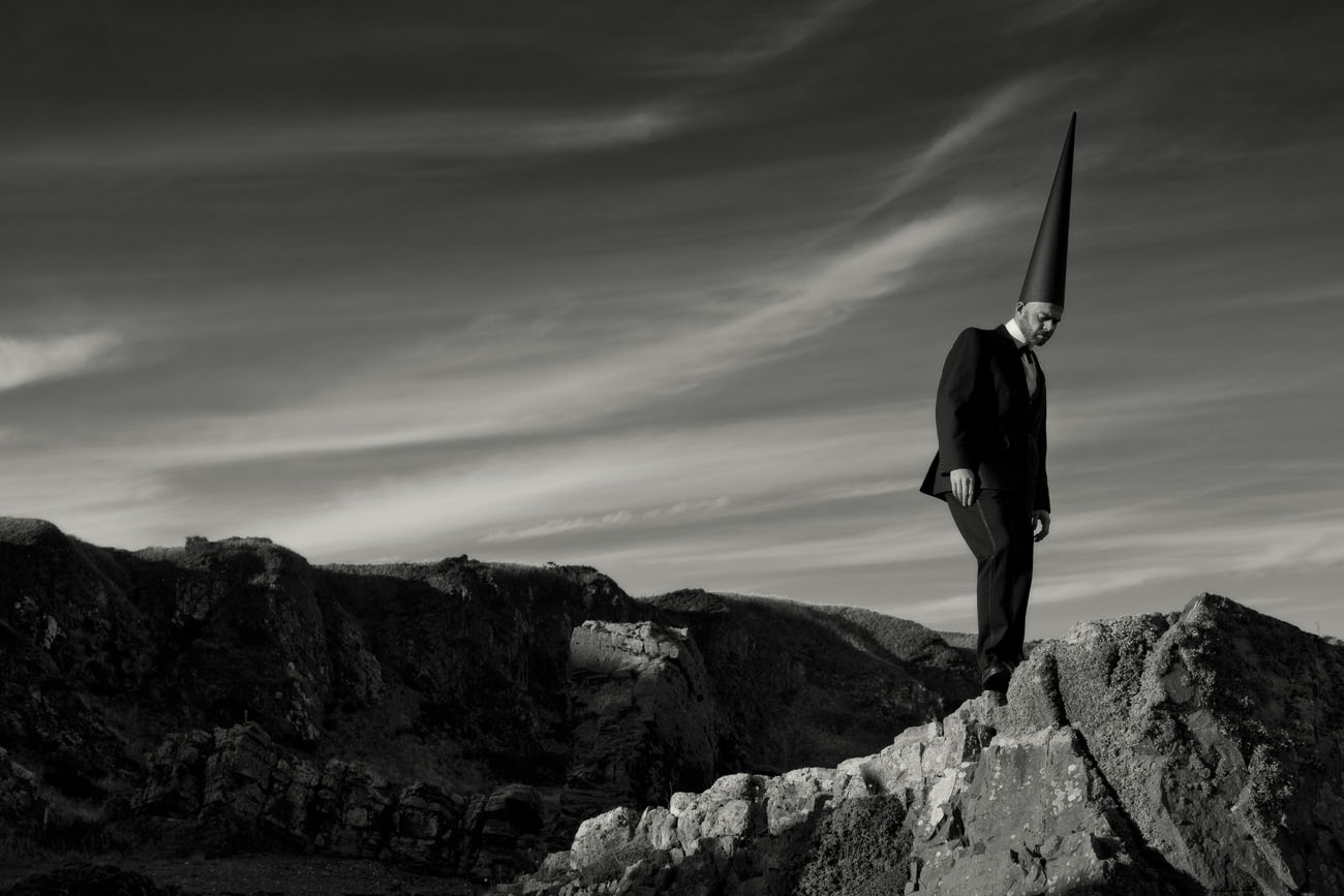 A lone coneman in a black suit and black pointed hat stands on a rocky outcrop with beautiful cirrus clouds in the sky behind. Black and white landscape photograph.
