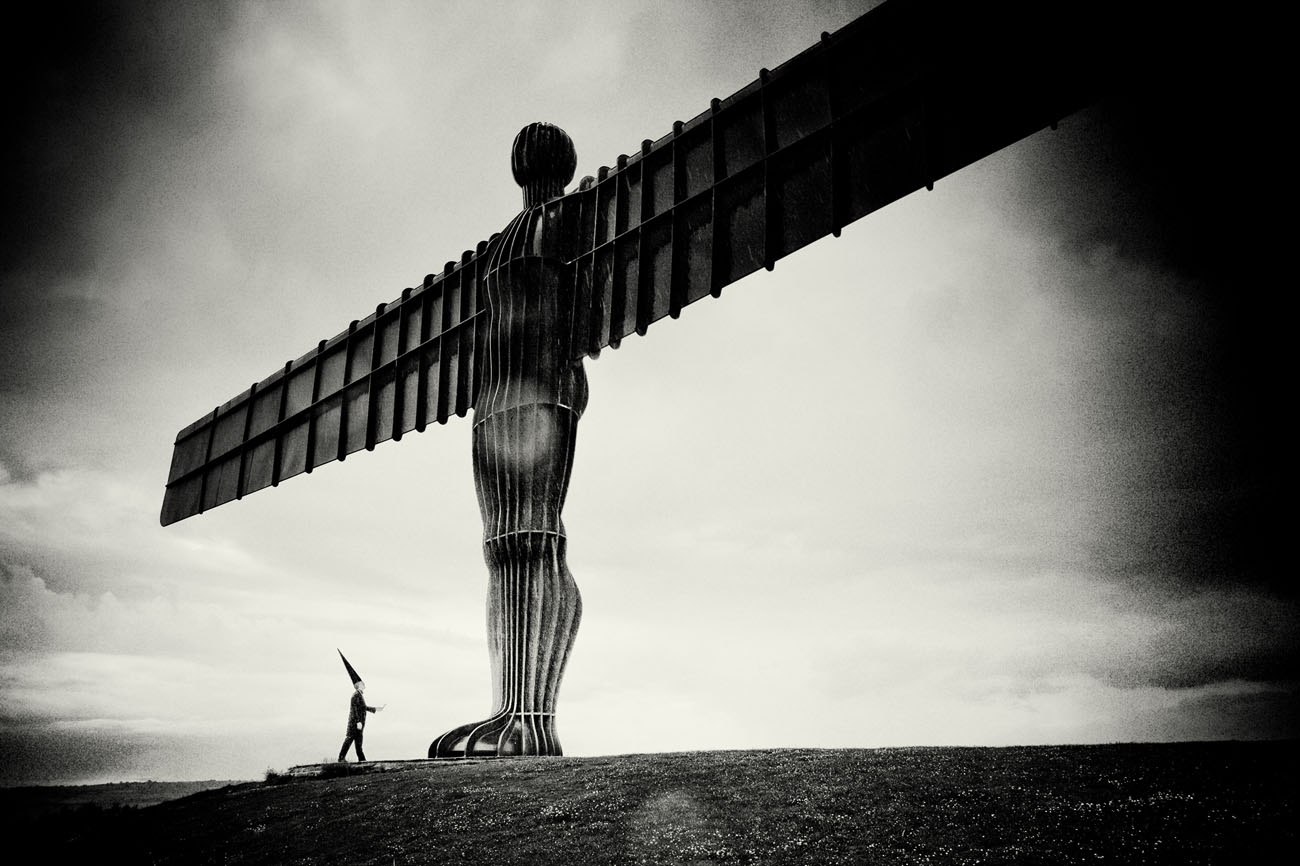 A coneman stands at the foot of the angel of the north with a feather in his hand. It's a stormy day and the photograph is black and white.