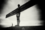 A coneman stands at the foot of the angel of the north with a feather in his hand. It's a stormy day and the photograph is black and white.