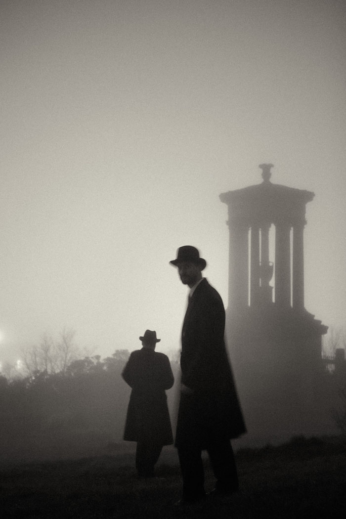 two men in long coats and hats in the mist onCalton Hill at night