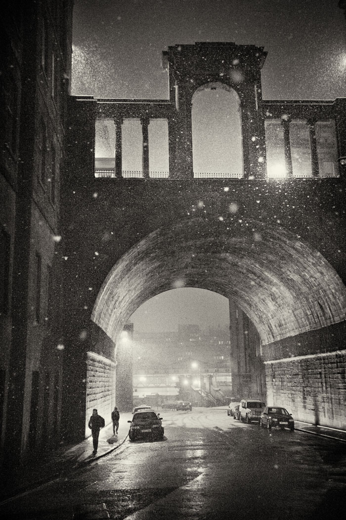 figures walk beneath Regent bridge in the snow at night.