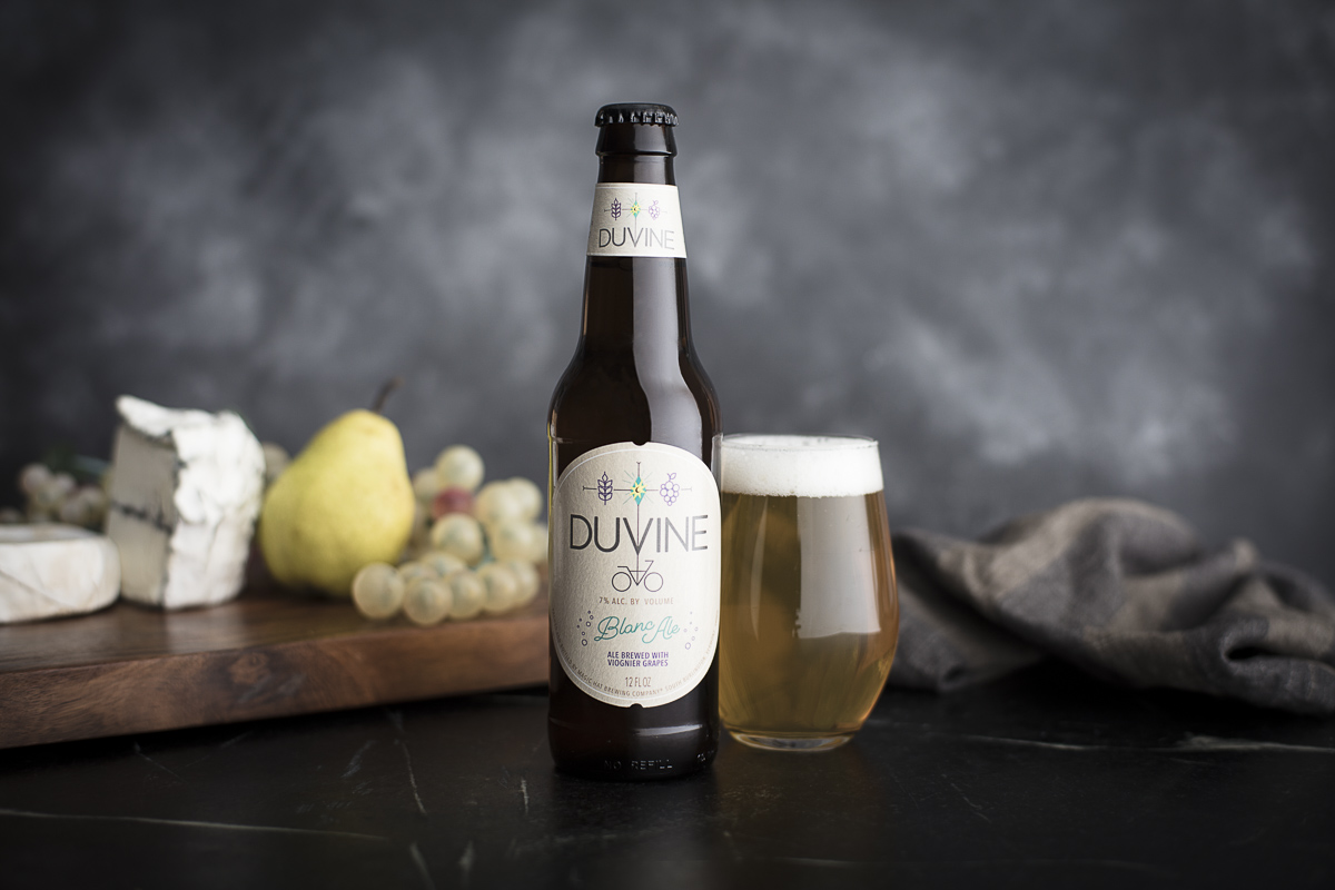 Magic Hat Brewing beer Duvine Blanc Ale on tabletop with a glass, pear, grapes and cheeses.
