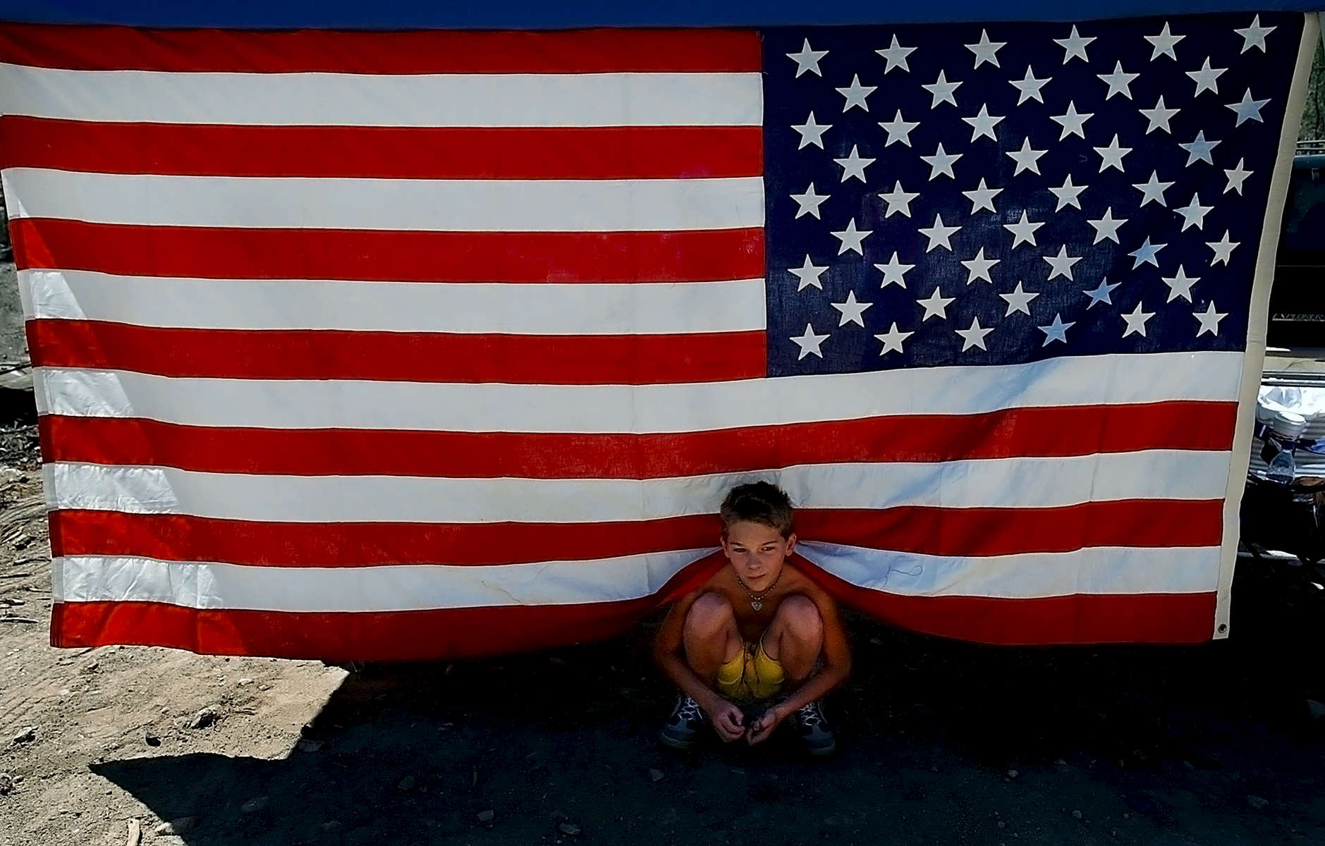 Jacob Burcham, 10, finds shade under an Americanflag that hangs from a Pop-Up shelter in the dirt and ash where his grandparents house was on Hook CreekRoad in Cedar Glen, Calif. The house was destroyed in the San Bernardino Old Fire last year. His grandparents, Janet and Jim Burcham, bought the house one week before the Old Fire burned through Cedar Glen. The Burchams were participating the Cedar Glen block partya year after the fire destroyed hundreds of homes. (The Press-Enterprise/Mark Zaleski)