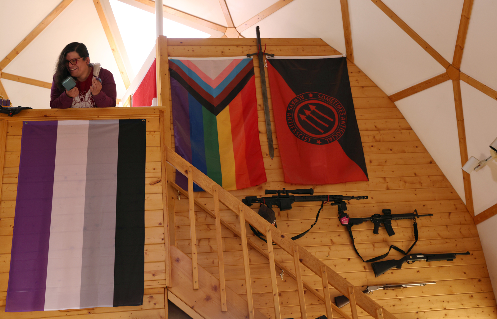 Bonnie Nelson chats with people as Nelson stands above the asexual flag as it hangs with several other flags representing gender and sexual identity at the Tenacious Unicorn Ranch in Westcliffe, Colorado. Above four mounted rifles, a red-and-black flag can be seen that says, “Sometimes antisocial, always antifascist,” with the three arrows of the Iron Front, a World War II-era German paramilitary anti-Nazi, antifascist organization. 
