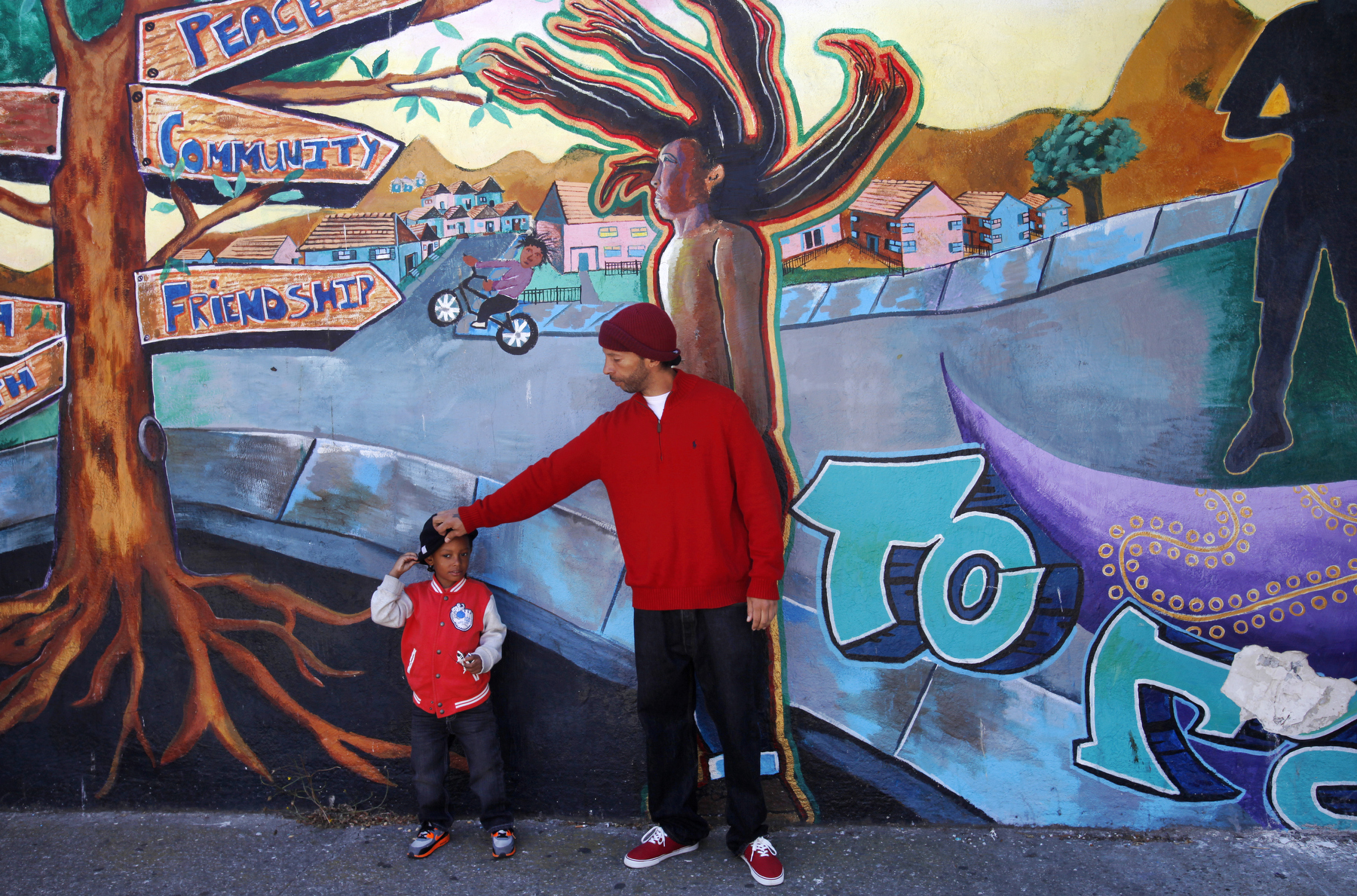 Sekou Carson adjusts the hat on his son Zakai Carson, 4, as they lean up against a community mural titled {quote}How Do You Shine{quote} while the two were out for a walk during Sekou\'s visit to the neighborhood to see his son July 3, 2014 in the Visitacion Valley neighborhood in San Francisco, Calif. {quote}My heart goes out to him and his family,{quote} Sekou said of the shooting, adding that he didn't feel the neighborhood was more unsafe now. {quote}I think it was an isolated incident,{quote} he said. Last Friday, June 27, outreach counselor Allen Calloway was shot and killed while playing dodgeball with children at a basketball court near Herz playground in Visitacion Valley. Since then, the summer program held at the playground has been closed temporarily and the area has had much fewer children and residents using it.