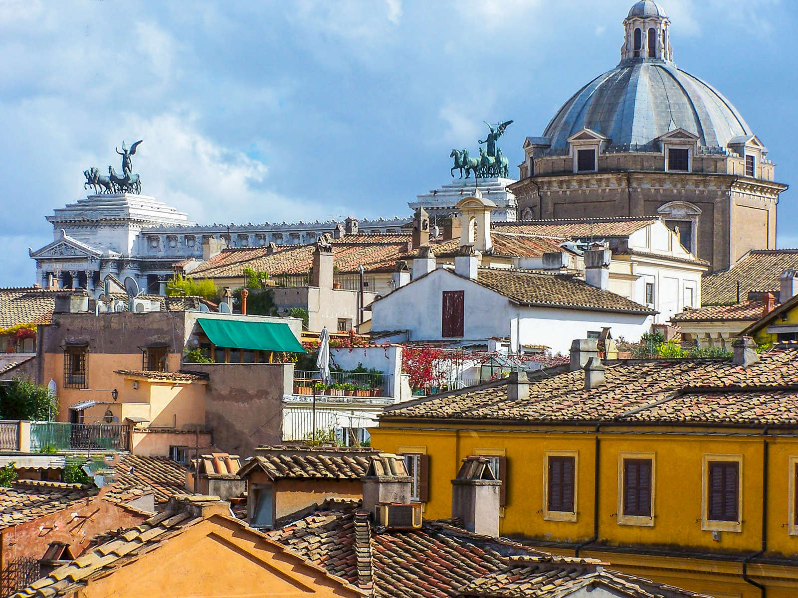 This image is a view overlooking the top of the National Monument to Victor Emmanuel II, and a lovely church dome peeking above the rooftops in Rome, Italy.  Prominently shown are bronze statues of the goddess Victory riding quadriga- detail on the roof of the monument.  The monument, also known as Altare della Patria, ({quote}Altar of the Fatherland{quote}), was built in honor of Victor Emmanuel, the first king of a unified Italy. The monument is also glaringly white, built from {quote}corpse-white marble{quote} imported from Botticino in Brescia, making it highly conspicuous amidst the generally brownish buildings surrounding it.  It occupies a site between the Piazza Venezia and the Capitoline Hill.  The monument is the largest in Rome.