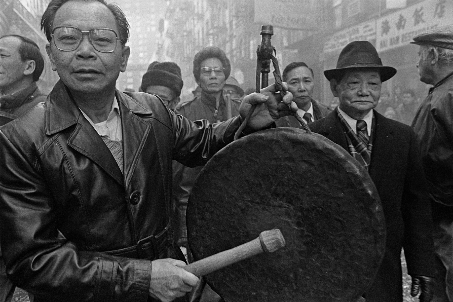 A group of men on Bayard St. during a Lunar New Year celebration in New York Chinatown in 1984. In the foreground a man is beating on a large gong.