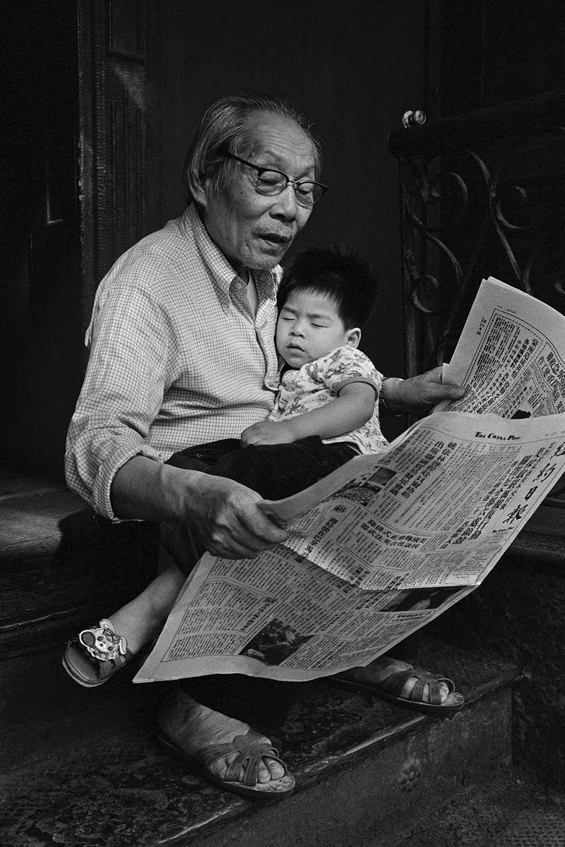 Kam Ho Lee, an elderly Chinese man, sits on the stoop of his apartment building, reading the newspaper, with his gandson, Vincent Lee, sleeping in his lap.