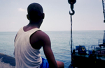 A boy sits on a ledge along the Malecon, watching men working and scuba diving from a barge. Havana, CUba