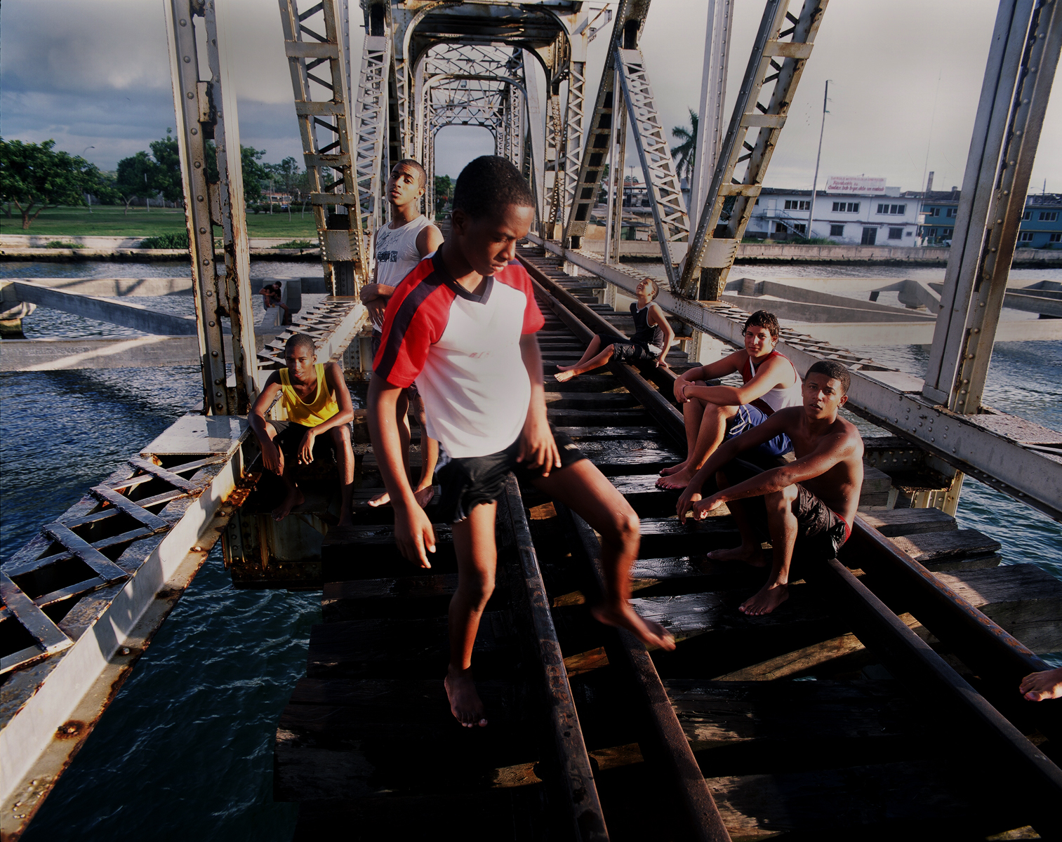 A teenage boy adjusts himself, while the other boys sit and talk, while resting or wating for their turn to jump from the railway bridge into waterway below. Both the railway bridge and waterway are used for commerical purposes by freight trains and commercial cargo ships. These teenage boys jump and swim at their own risk due to a lack of laws, police enforcement, or opportunites for jobs or educational summer camps.Mantanzas, Cuba