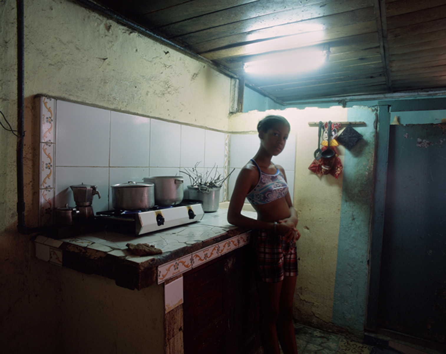 Nazarena, prepares dinner,--chicken, rice, and black beans--in a makeshift kitchen, with dripping water, a hotplate, and two pots. Despite her stark surroundings and limited resources, she is always smiling and listening to music whiling preparing a feast. Havana, Cuba