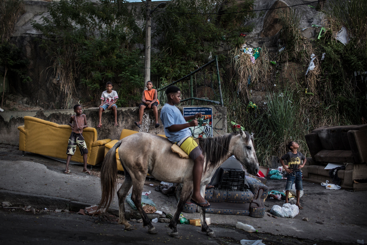 A teenager rides a horse down the street in the Complexo do Penha,behind him, some of his friends spend the afternoon playing with kites, in Rio de Janeiro, Brazil