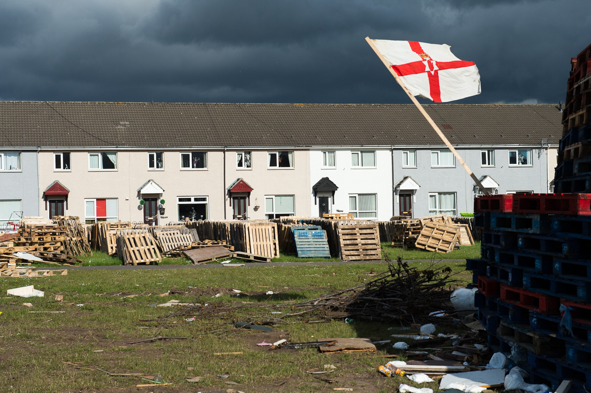 The Ulster Banner, the former flag of the Northern Ireland government (1953–1972), is seen on a bonfire-under-construction in the Protestant Lower Shankill area of West Belfast. The flag no longer has an official status but has become a contentious symbol representing Ulster loyalism.Bonfires are traditionally lit in Protestant areas on the {quote}Eleventh Night{quote} of July to commemorate the victory of the Protestant William of Orange over his Catholic father-in-law King James II at the Battle of the Boyne in 1690. Preparations start months before with the collection of pallets and other materials like rubber tires. The bonfire is followed on July 12 by a large parade held by the Orange Order — a Protestant fraternal organization —  and loyalist marching bands. The {quote}Twelfth{quote} as it is called is a tense period. Many Catholics consider the celebrations a display of sectarian triumphalism whereas for many working-class Protestants they are an essential expression of their cultural identity.