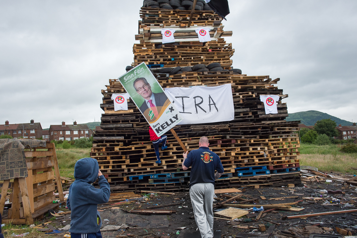 A young man wearing a uniform shirt of the Pride of Ardoyne Flute Band  — one of the many loyalist bands in Belfast, some of which are linked to paramilitary groups — adds an election poster of a Sinn Féin politician to a finished bonfire in the Protestant Glenbryn area of north Belfast. Other hated symbols on the bonfire are a banner referring to the IRA and signs protesting  restrictions on marching parades by the Parades Commission -an official bipartisan parade watchdog.Bonfires are traditionally lit in Protestant areas on the {quote}Eleventh Night{quote} of July to commemorate the victory of the Protestant William of Orange over his Catholic father-in-law King James II at the Battle of the Boyne in 1690. Preparations start months before with the collection of pallets and other materials like rubber tires. The bonfire is followed on July 12 by a large parade held by the Orange Order — a Protestant fraternal organization — and loyalist marching bands. The {quote}Twelfth{quote} as it is called is a tense period. Many Catholics consider the celebrations a display of sectarian triumphalism whereas for many working-class Protestants they are an essential expression of their cultural identity.