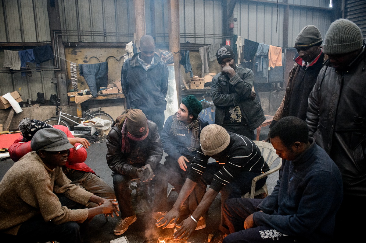 Calais, December 2014 Sudanese migrants warm themselves over a fire in the cold, damp and smoke-filled Galoo squat, a large abandoned metal recycling plant.
