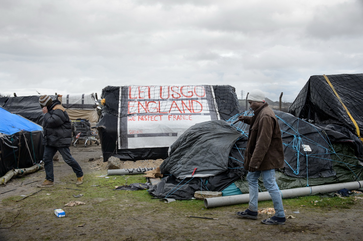 Calais, December 2014 Migrants walk by shelters made of plastic sheeting, packing crates and other found materials in Tioxide camp in Calais. The makeshift tent and tarpaulin camps are locally known as {quote}jungles{quote}.  The Tioxide jungle, situated next to a chemical plant, is currently the largest camp in Calais.